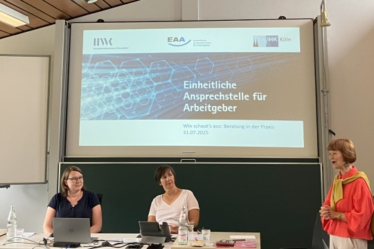 Auf dem Bild sieht man zwei Personen, die vor einer PowerPoint Präsentation sitzen. Rechts von ihnen steht eine weitere Person, die spricht. Auf der Folie der Präsentation sind die Logos der EAA, der HWK und der IHK zu sehen sowie der Titel: Einheitliche Ansprechstellen für Arbeitgeber - Wie schauts aus: Beratung in der Praxis.