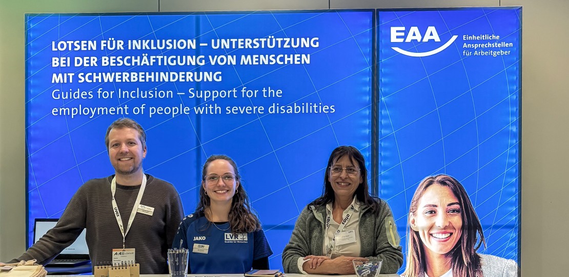 Auf dem Bild sind drei Fachberatende an dem EAA-Infostand zu sehen. Sie stehen vor einer großen blauen Rückwand, auf der Lotsen für Inklusion steht, und hinter eine Infotheke, auf der Flyer un Werbemittel ausgelegt sind.