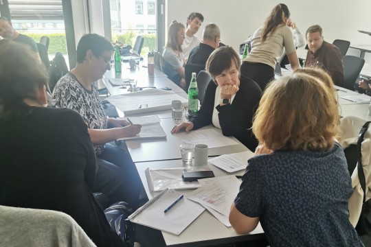 Auf dem Bild sind zwei Gruppen von EAA-Fachberatenden zu sehen, die mit Stiften und Zetteln an Tischen in einem Besprechungsraum sitzen und miteinander reden