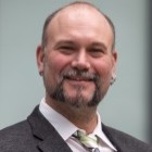 Potrait von Herrn Sybertz. Er hat sehr kurze graue Haare und einen kurzen Bart. Er trägt ein weißes Hemd mit grauem Jacket.